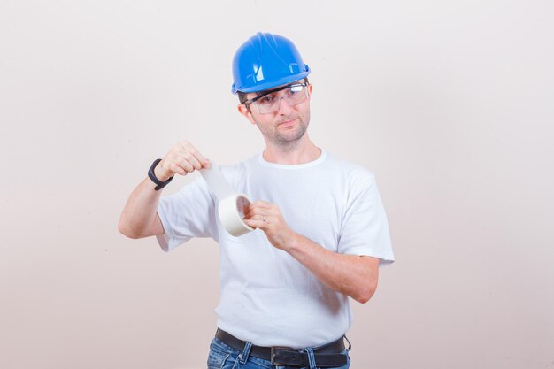 Молодой человек открывает рулон изоленты в футболке, джинсах, шлеме и смотрит внимательно