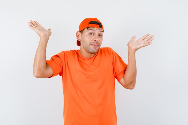 Young man in orange t-shirt and cap raising hands in helpless manner and looking cute