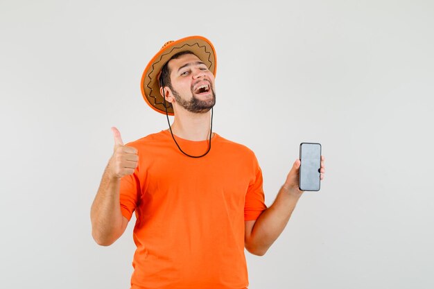 Young man in orange t-shirt, hat holding mobile phone with thumb up and looking glad , front view.