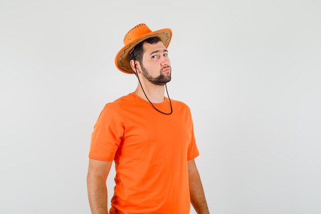 Молодой человек в оранжевой футболке, шляпе смотрит и выглядит уверенно, вид спереди.