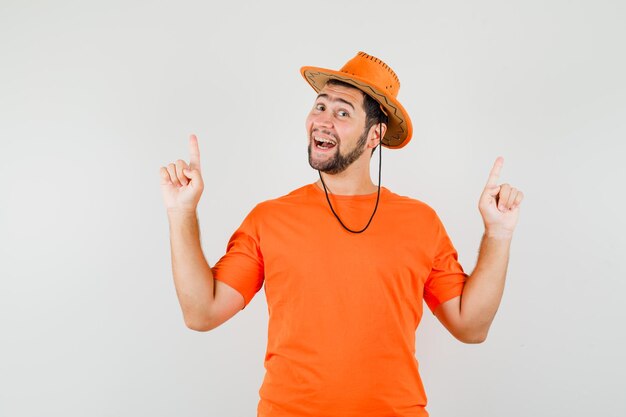 Young man in orange t-shirt, hat pointing fingers up and looking optimistic , front view.