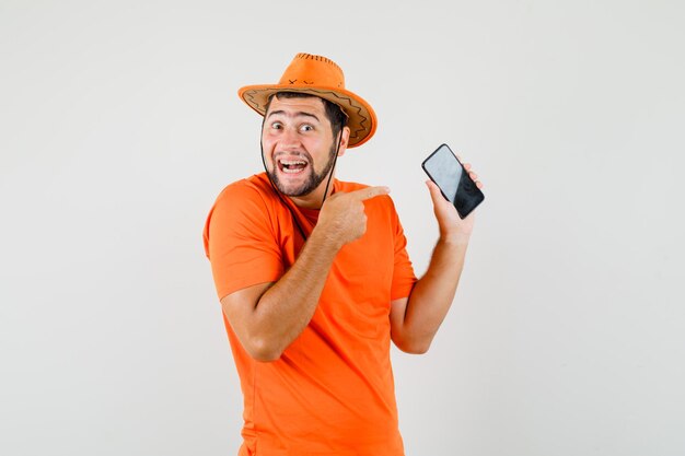 주황색 티셔츠를 입은 젊은 남자, 모자는 휴대전화를 가리키며 쾌활하고 앞모습을 보고 있습니다.