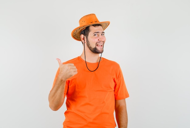 Young man in orange t-shirt, hat showing thumb up and looking glad , front view.