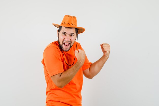 주황색 티셔츠를 입은 청년, 우승자 제스처를 보여주고 행복해 보이는 모자, 앞모습.