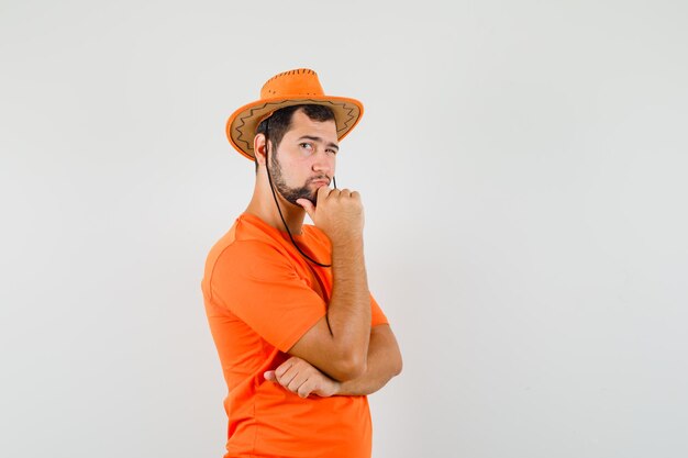 オレンジ色のTシャツを着た若い男、思考ポーズで立って、賢明に見える帽子、正面図。