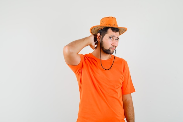 Young man in orange t-shirt, hat touching his neck with fingers and looking handsome , front view.