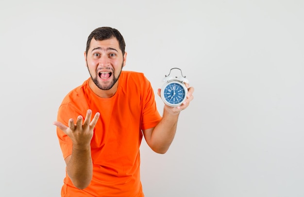 Free photo young man in orange t-shirt holding alarm clock and screaming , front view.