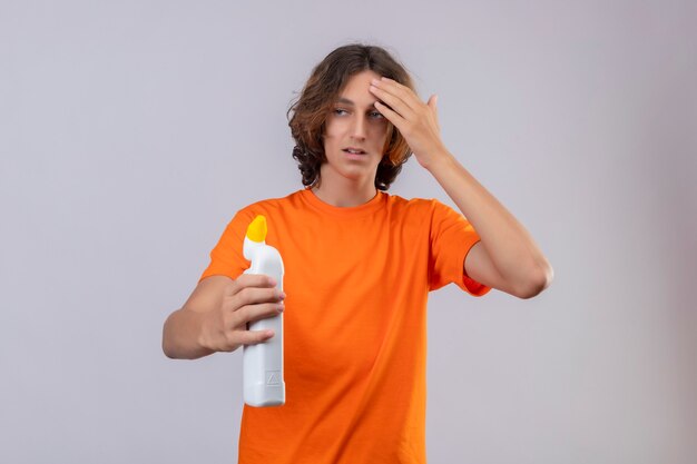 白い背景の上に混乱して驚いて立っている探しているクリーニング用品のボトルを保持しているオレンジ色のtシャツの若い男