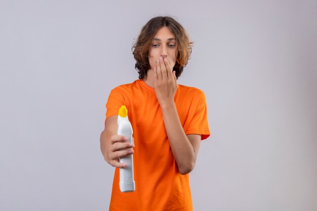 Young man in orange t-shirt holding bottle of cleaning supplies looking at it surprised and amazed covering mouth with hand standing over white background