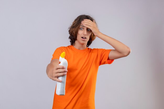 Young man in orange t-shirt holding bottle of cleaning supplies looking at it surprised touching head with hand standing over white background
