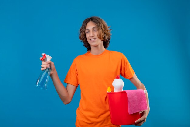 Young man in orange t-shirt  holding bucket with cleaning tools and cleaning spray looking at camera with confident smile on face standing over blue background