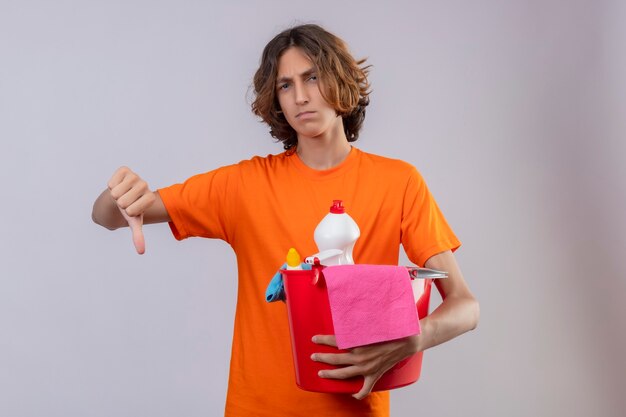 Young man in orange t-shirt holding bucket with cleaning tools looking at camera displeased showing thumbs down standing over white background