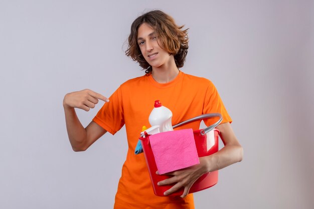 Young man in orange t-shirt holding bucket with cleaning tools pointing with finger to it looking at camera smiling cheerfully standing over white background