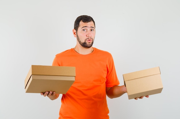 Young man in orange t-shirt holding cardboard boxes and looking confused , front view.