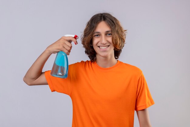 Young man in orange t-shirt  holding cleaning spray looking at camera smiling cheerfully standing over white background