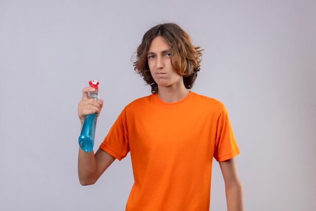 Young man in orange t-shirt  holding cleaning spray looking at camera with sad expression on face standing over white background