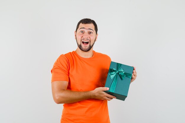 Young man in orange t-shirt holding present box and looking glad , front view.