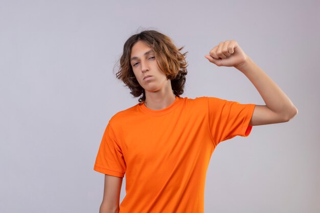 Молодой человек в оранжевой футболке смотрит в камеру с уверенным выражением лица, поднимая кулак, самодовольный победитель, стоящий на белом фоне