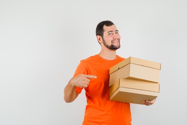 Young man in orange t-shirt pointing at cardboard boxes and looking happy , front view.