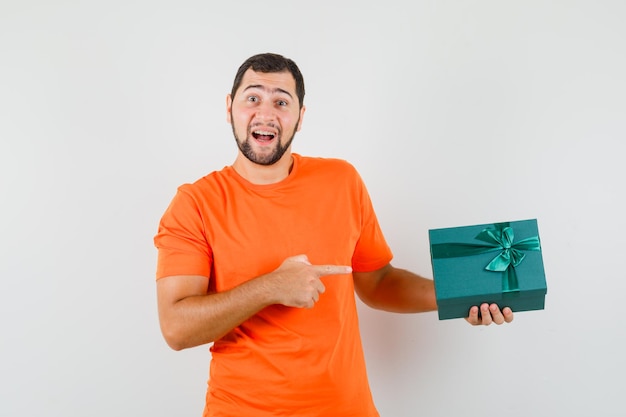 Young man in orange t-shirt pointing at present box and looking glad , front view.