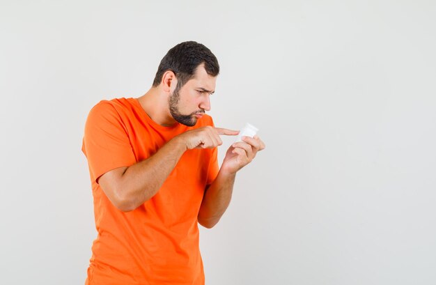 Молодой человек в оранжевой футболке, читая информацию о бутылке таблеток, вид спереди.