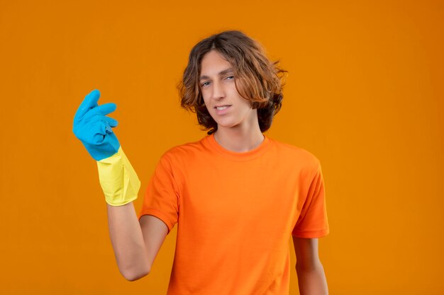 Молодой человек в оранжевой футболке в резиновых перчатках жестикулирует рукой, делая денежный жест, глядя в камеру с уверенной улыбкой, стоя на желтом фоне