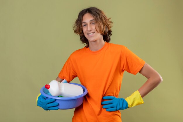 Молодой человек в оранжевой футболке в резиновых перчатках держит таз с чистящими средствами, глядя в камеру, улыбаясь позитивно и счастливо стоя на зеленом фоне
