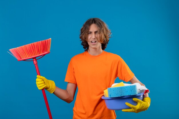 Young man in orange t-shirt wearing rubber gloves holding basin with cleaning tools and mop looking annoyed standing over blue background