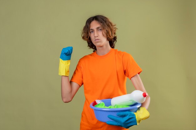 Young man in orange t-shirt wearing rubber gloves holding basin with cleaning tools raising fist threatening with frowning face standing over green background