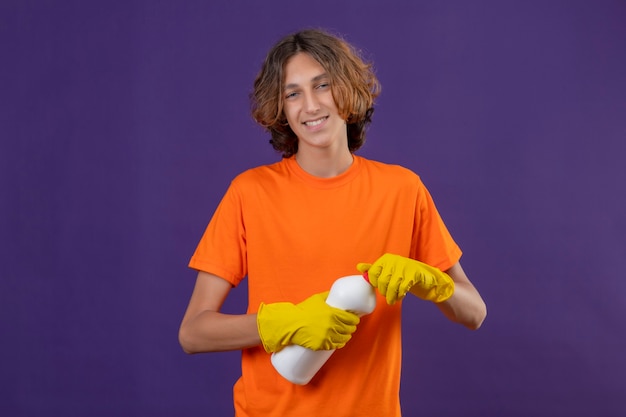 보라색 배경 위에 유쾌하게 서있는 카메라를보고 청소 용품 병을 들고 고무 장갑을 끼고 오렌지 티셔츠에 젊은 남자
