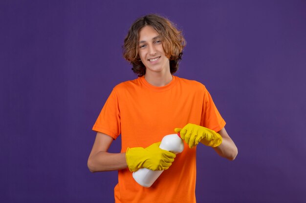 Young man in orange t-shirt wearing rubber gloves holding bottle of cleaning supplies looking at camera smiling cheerfully standing over purple background