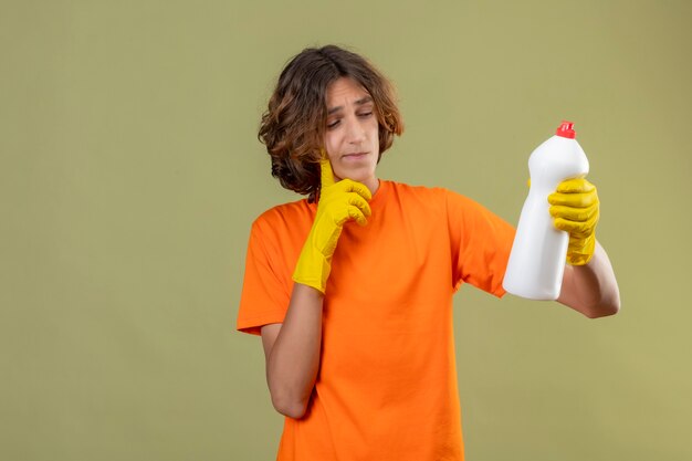 Молодой человек в оранжевой футболке в резиновых перчатках держит бутылку чистящих средств, глядя на нее с задумчивым выражением лица, думая, стоя на зеленом фоне