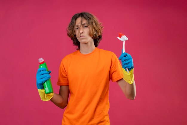 Young man in orange t-shirt wearing rubber gloves holding bottle of cleaning supplies and scrubbing brush looking at camera displeased standing over pink background