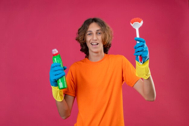 Young man in orange t-shirt wearing rubber gloves holding bottle of cleaning supplies and scrubbing brush looking at camera smiling cheerfully happy and positive standing over pink backgroun