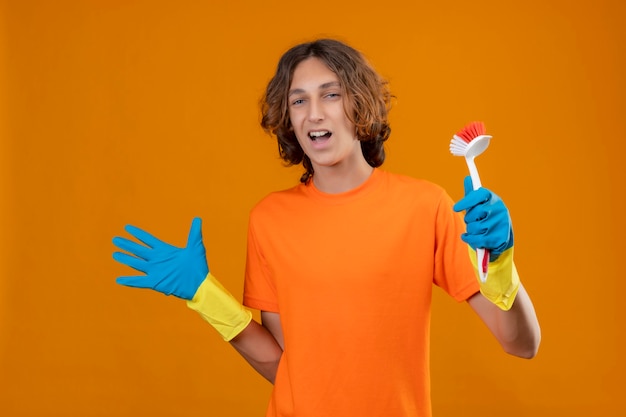 Young man in orange t-shirt wearing rubber gloves holding bottle of cleaning supplies standing with arm raised smiling cheerfully  happy and positive standing over yellow background