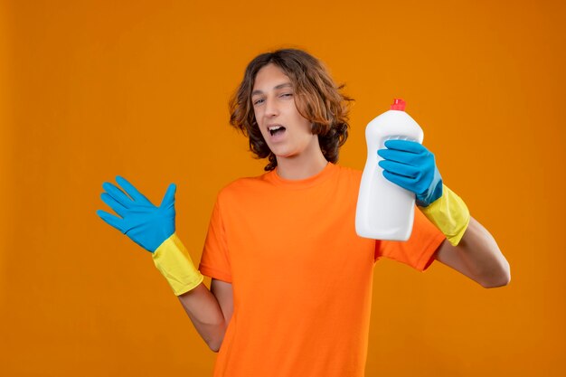 Молодой человек в оранжевой футболке в резиновых перчатках держит бутылку чистящих средств, стоя с поднятой рукой, весело улыбаясь и позитивно стоит на желтом фоне
