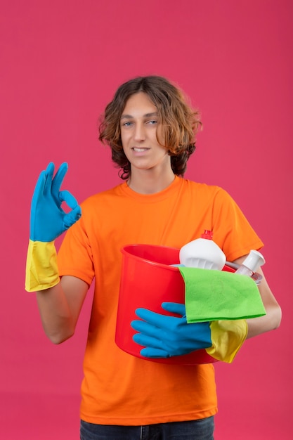 Young man in orange t-shirt wearing rubber gloves holding bucket with cleaning tools looking at camera smiling confident doing ok sign standing over pink background