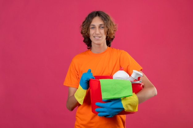 Молодой человек в оранжевой футболке в резиновых перчатках держит ведро с чистящими средствами и смотрит в камеру, улыбаясь позитивно и счастливо стоя на розовом фоне