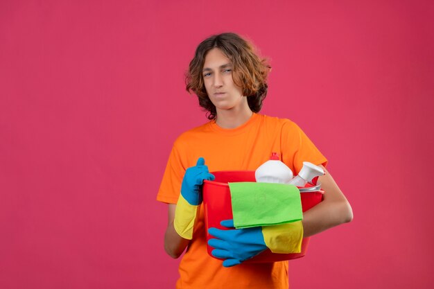 분홍색 배경 위에 서있는 얼굴에 회의적인 표정으로 카메라를보고 청소 도구와 양동이를 들고 고무 장갑을 끼고 오렌지 티셔츠에 젊은 남자