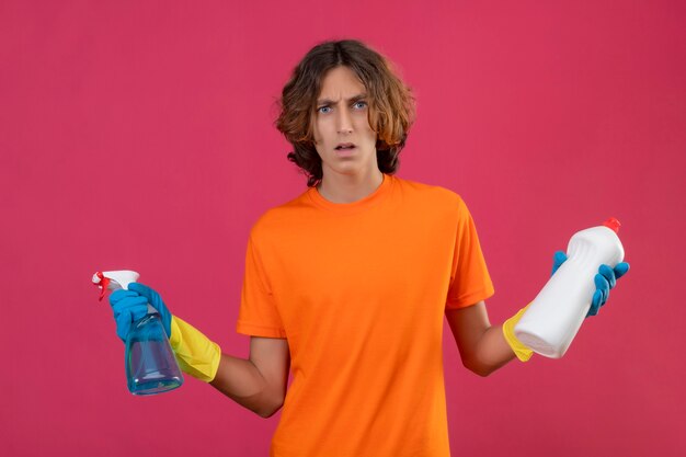 분홍색 배경 위에 서있는 카메라를보고 혼란 스 러 워 확산 손을 찾고 청소 스프레이 및 청소 용품 병을 들고 고무 장갑을 끼고 오렌지 티셔츠에 젊은 남자