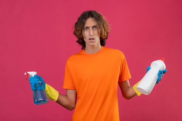 Young man in orange t-shirt wearing rubber gloves holding cleaning spray and bottle of cleaning supplies looking at camera confused spreading hands standing over pink background