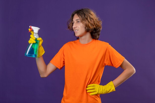 Young man in orange t-shirt wearing rubber gloves holding cleaning spray  looking aside  smiling with happy face  ready to clean standing over purple background