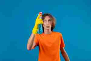 Free photo young man in orange t-shirt wearing rubber gloves holding cleaning spray looking up with pensive expression on face standing over blue background