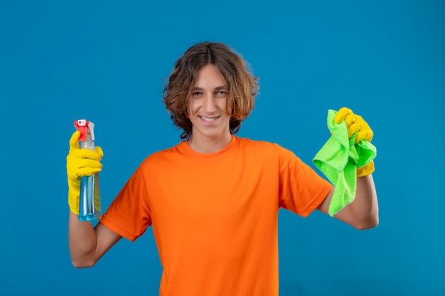 Young man in orange t-shirt wearing rubber gloves holding cleaning spray and rug looking at camera with confident smile ready to clean standing over blue background