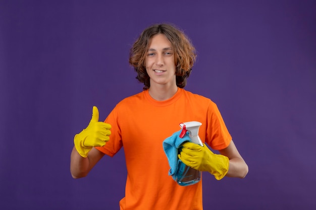 Young man in orange t-shirt wearing rubber gloves holding cleaning spray and rug looking at camera with confident smile showing thumbs up ready to clean standing over purple background