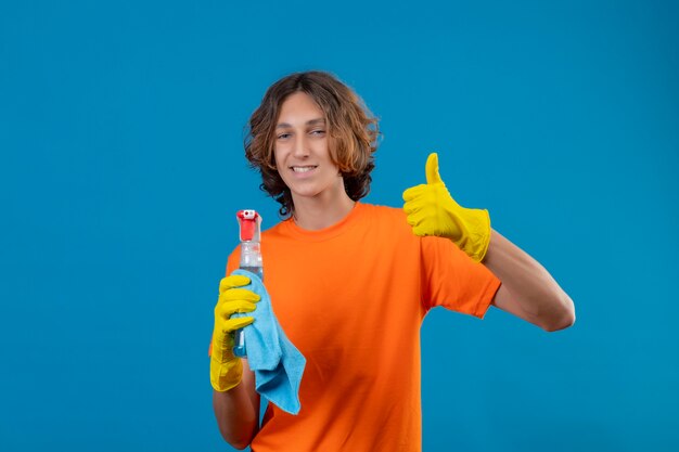 Young man in orange t-shirt wearing rubber gloves holding cleaning spray and rug looking at camera with confident smile showing thumbs up standing over blue background
