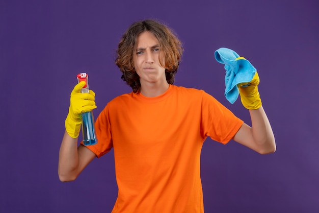 オレンジ色のtシャツを着た若い男がクリーニングスプレーと敷物を保持している紫色の背景の上に立っている顔に懐疑的な表情でカメラを見てゴム手袋を着用