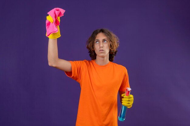 Young man in orange t-shirt wearing rubber gloves holding cleaning spray and rug looking up displeased ready to clean standing over purple background
