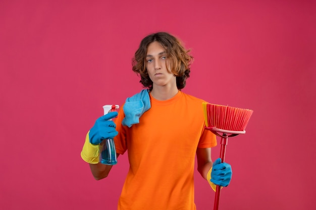 Молодой человек в оранжевой футболке в резиновых перчатках держит швабру и чистящий спрей, смотрит в камеру с серьезным и уверенным выражением лица, стоя на розовом фоне