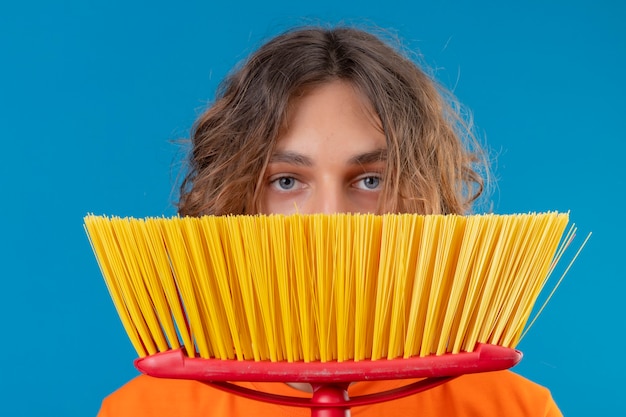 Free photo young man in orange t-shirt wearing rubber gloves holding mop  hiding behind it standing over blue background